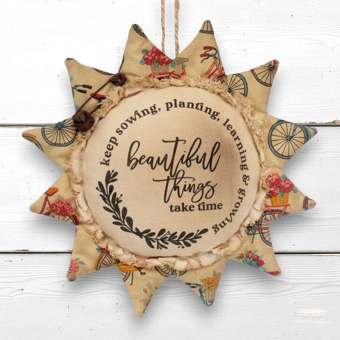 A fabric sunflower-shaped hanging ornament with a rustic twine loop at the top, featuring a central beige circle with the phrase 'beautiful things take time' and petals made from floral and bicycle-patterned fabric, shown against a white wood plank background.