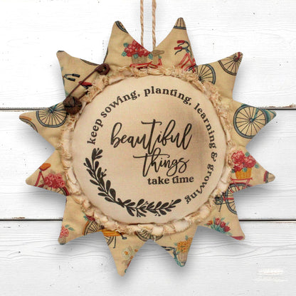 A fabric sunflower-shaped hanging ornament with a rustic twine loop at the top, featuring a central beige circle with the phrase 'beautiful things take time' and petals made from floral and bicycle-patterned fabric, shown against a white wood plank background.
