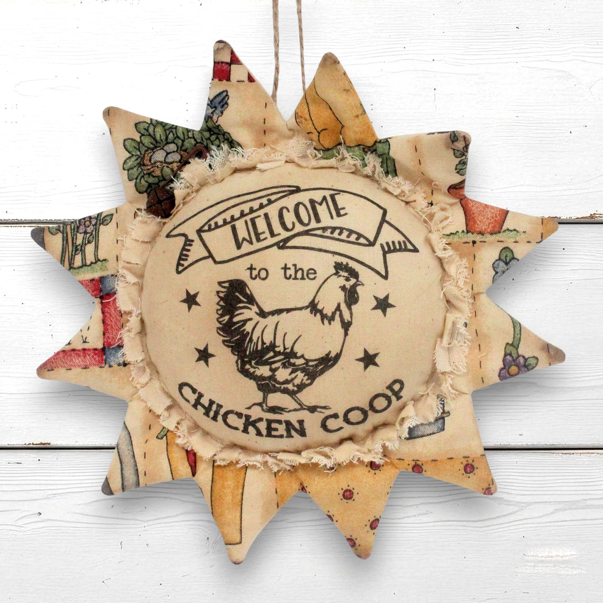 A rustic fabric sunflower ornament hanging with a central circular patch that reads “Welcome to the Chicken Coop” and shows a rooster illustration. The outer rays have patchwork floral and botanical prints in muted tones, and the piece hangs against a white wood plank background.
