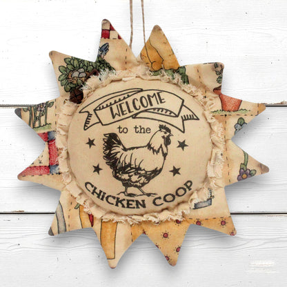 A rustic fabric sunflower ornament hanging with a central circular patch that reads “Welcome to the Chicken Coop” and shows a rooster illustration. The outer rays have patchwork floral and botanical prints in muted tones, and the piece hangs against a white wood plank background.