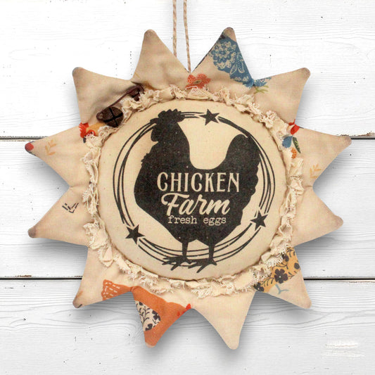 A fabric sunflower-shaped hanging ornament with a central round panel reading “Chicken Farm Fresh eggs” and a black chicken silhouette, surrounded by petals on a white wooden background
