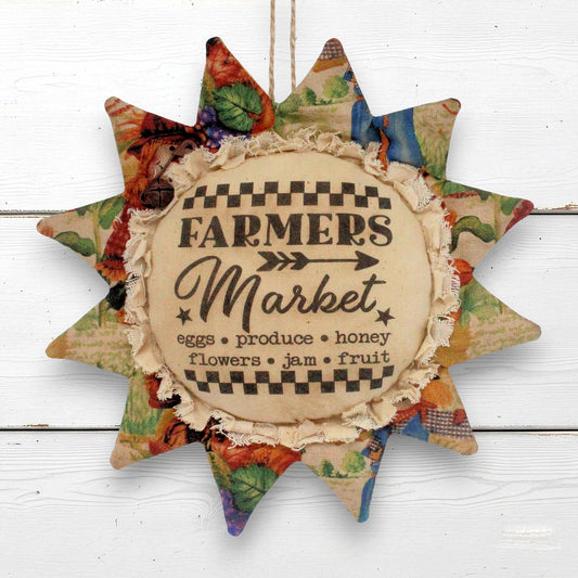 A rustic fabric sunflower ornament with a center reading “FARMERS Market” and multicolored patchwork fabric rays around the edge, shown against a white wood background.