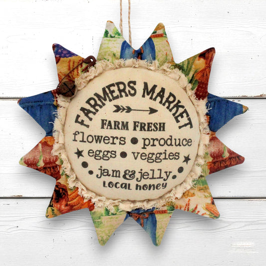 A rustic sunflower-shaped hanging ornament with a central round label reading “Farmers Market” and “Farm Fresh flowers produce eggs veggies jam & jelly local honey,” surrounded by multicolored patterned fabric petals on a white wood background.