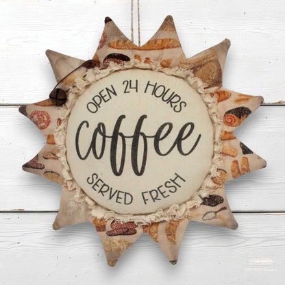 A decorative sunflower ornament wall hanging with a rustic fabric design, featuring the words 'OPEN 24 HOURS coffee SERVED FRESH' on a light beige center panel, shown hanging against a white wood plank background.