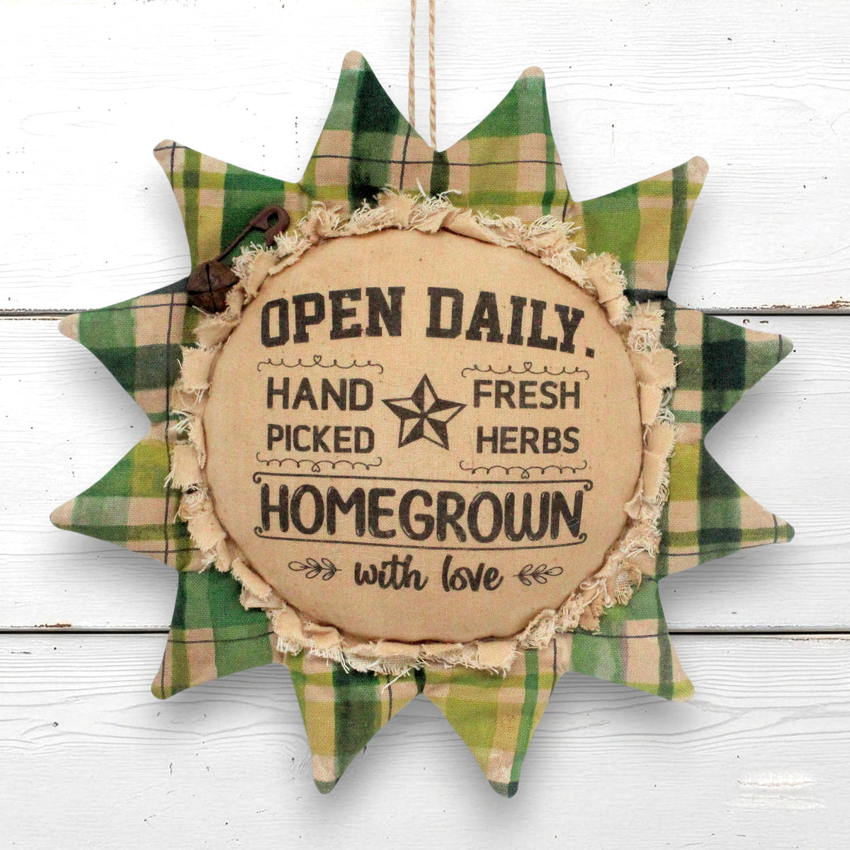 A rustic fabric sunflower-shaped hanging ornament with green and beige plaid petals, a frayed textured center, and printed text reading “OPEN DAILY. HAND PICKED FRESH HERBS HOMEGROWN with love,” shown against a white wood plank background.