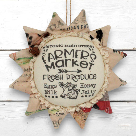 A rustic fabric sunflower hanging ornament with a faux-rustic 'Farmers Market' sign in the center, surrounded by patterned starburst points, hanging against a white wood plank background.