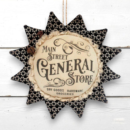 A decorative sunflower-shaped wall hanging with a central vintage-style sign reading 'Main & Street General Store' and 'Dry Goods Hardware Groceries,' surrounded by black fabric petals with a small white floral pattern on a white wood plank background.
