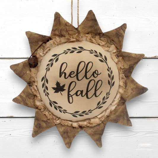 A decorative autumn sunflower-shaped  ornament that reads 'hello fall' in black script with a leaf motif. It hangs against a white wood plank background.