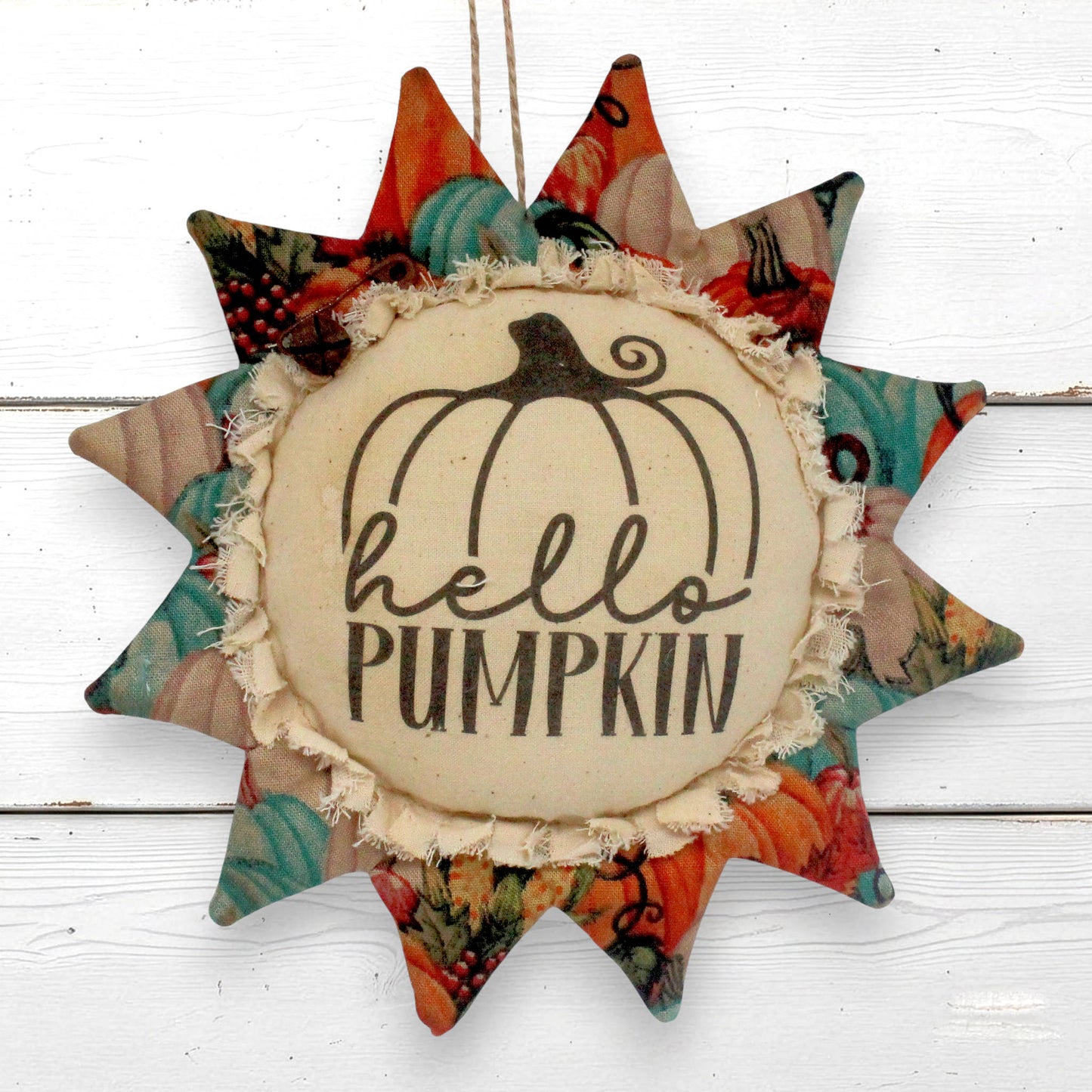 A fabric sunflower ornament hanging with a central cream circle reading 'hello PUMPKIN' and a black pumpkin outline, surrounded by patterned autumn star points in orange, teal, beige, and red tones, photographed against a white wood background.