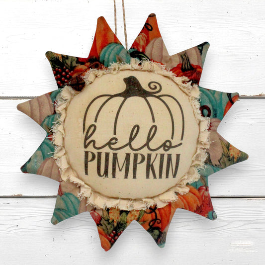 A fabric sunflower ornament hanging with a central cream circle reading 'hello PUMPKIN' and a black pumpkin outline, surrounded by patterned autumn star points in orange, teal, beige, and red tones, photographed against a white wood background.