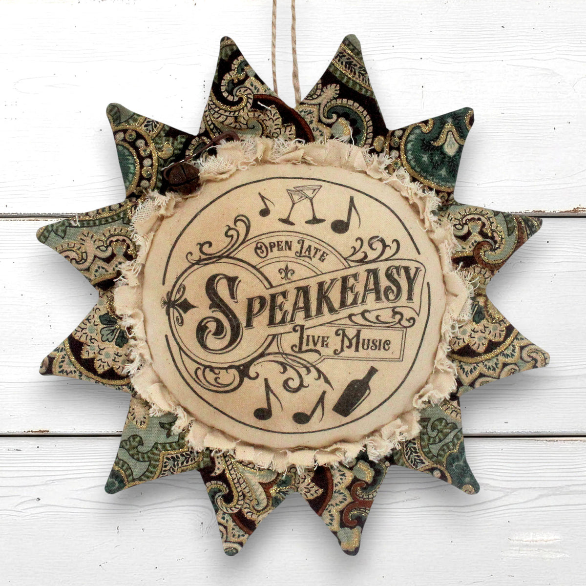 A decorative fabric sunflfower ornament wall hanging with a central circular patch that reads 'Open Late Speakeasy Live Music' with musical notes and a bottle illustration. The outer points feature vintage paisley patterns in muted green, brown, and beige tones, and the piece hangs against a white wood plank background.
