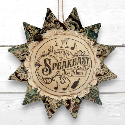 A decorative fabric sunflfower ornament wall hanging with a central circular patch that reads 'Open Late Speakeasy Live Music' with musical notes and a bottle illustration. The outer points feature vintage paisley patterns in muted green, brown, and beige tones, and the piece hangs against a white wood plank background.
