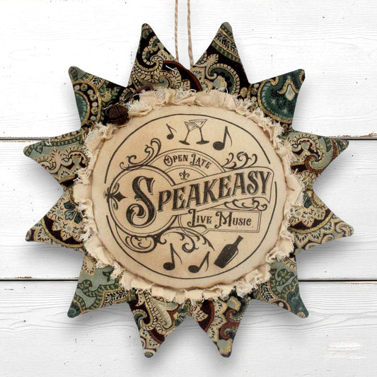 A decorative fabric sunflfower ornament wall hanging with a central circular patch that reads 'Open Late Speakeasy Live Music' with musical notes and a bottle illustration. The outer points feature vintage paisley patterns in muted green, brown, and beige tones, and the piece hangs against a white wood plank background.