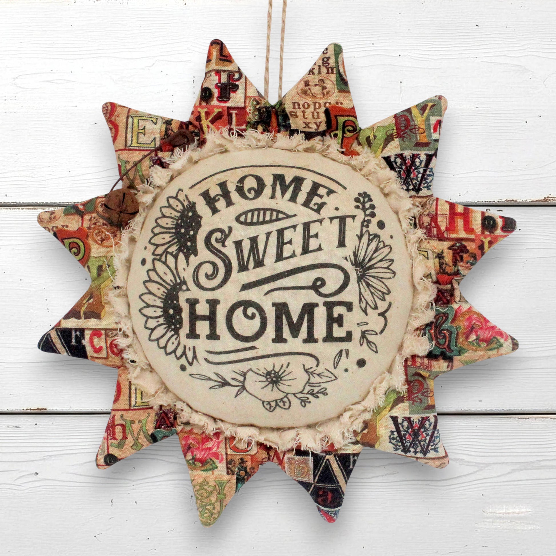 A handmade fabric sunflower-shaped wall hanging with the words 'HOME SWEET HOME' printed in the center, surrounded by a patchwork of vintage-style patterned paper and ruffled trim, hanging against a white wood plank background.