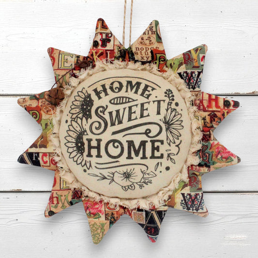 A handmade fabric sunflower-shaped wall hanging with the words 'HOME SWEET HOME' printed in the center, surrounded by a patchwork of vintage-style patterned paper and ruffled trim, hanging against a white wood plank background.