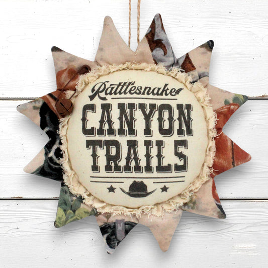 A rustic sunflower fabric hanging sign with the words 'Rattlesnaker CANYON TRAILS' in black lettering on a cream center panel, surrounded by camouflage and western-patterned points, shown against a white wood plank background.