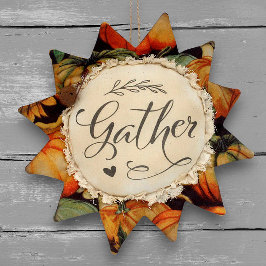 A fabric sunflower-shaped hanging ornament with a central cream circle reading “Gather” in black script, surrounded by autumn pumpkin-patterned petals, shown against a gray wood plank background.