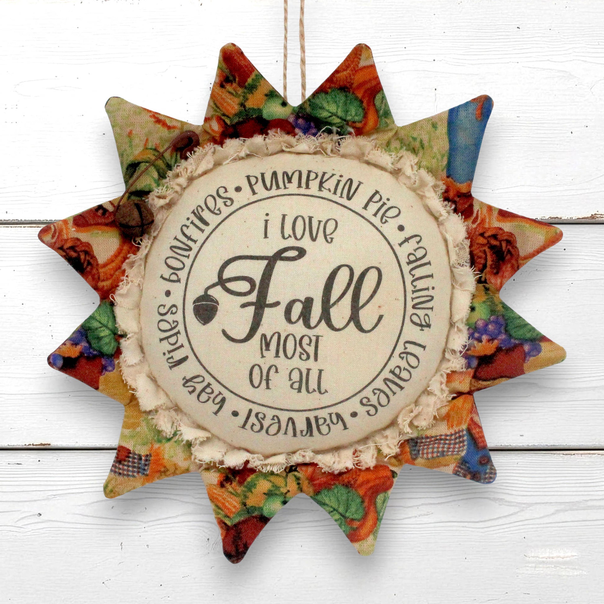 A fabric sunflower autumn hanging ornament with a central round sign that reads 'i love Fall most of all' and lists seasonal phrases around the edge. The star has multicolor fall-themed patterned fabric petals and a rustic beige fringed center, shown hanging against a white wood plank background.