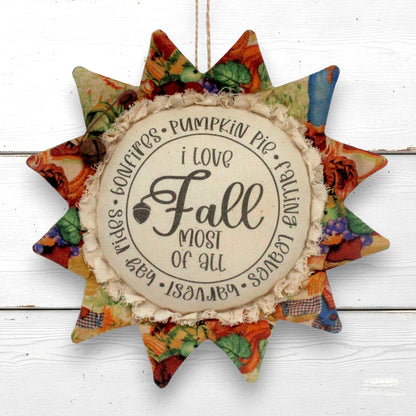 A fabric sunflower autumn hanging ornament with a central round sign that reads 'i love Fall most of all' and lists seasonal phrases around the edge. The star has multicolor fall-themed patterned fabric petals and a rustic beige fringed center, shown hanging against a white wood plank background.