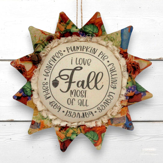 A fabric sunflower autumn hanging ornament with a central round sign that reads 'i love Fall most of all' and lists seasonal phrases around the edge. The star has multicolor fall-themed patterned fabric petals and a rustic beige fringed center, shown hanging against a white wood plank background.