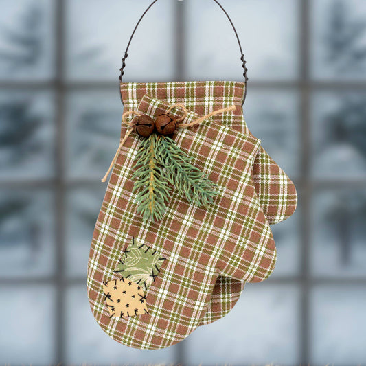 A brown and green plaid mitten-shaped hanging ornament with a black wire hanger, decorated with pinecones, a sprig of greenery, and a small pinecone patch on the front, set against a blurred window background.