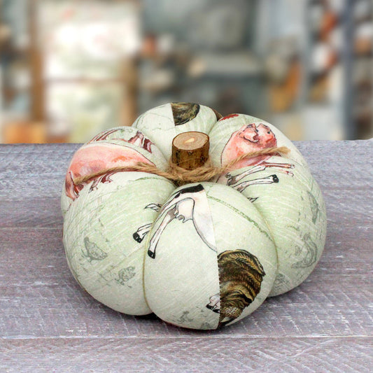 A decorative fabric pumpkin with a pale sage green base, printed with black line drawings of animals, sitting on a gray wooden surface with a blurred background.