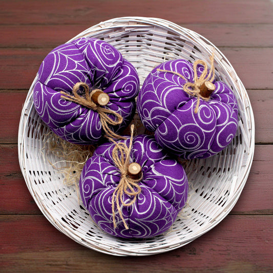 Three purple fabric pumpkins with decorative ribbons in a white woven basket on a wooden surface.