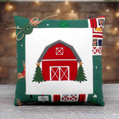 Small accent pillow with an embroidered design of a classic red barn with a Christmas tree on both sides, trimmed in a fabric with a barn and reindeer design