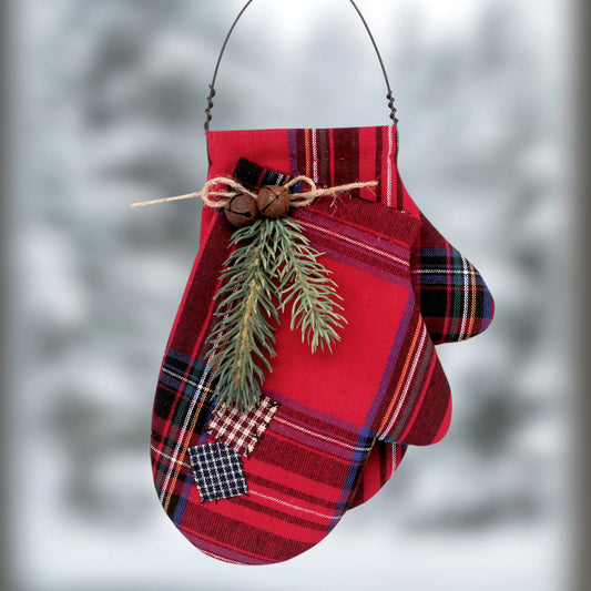 A red and black plaid fabric mitten ornament hanging from a black wire loop. It features a pine branch and two small wooden beads tied around the wrist area, set against a blurred, snowy outdoor background.