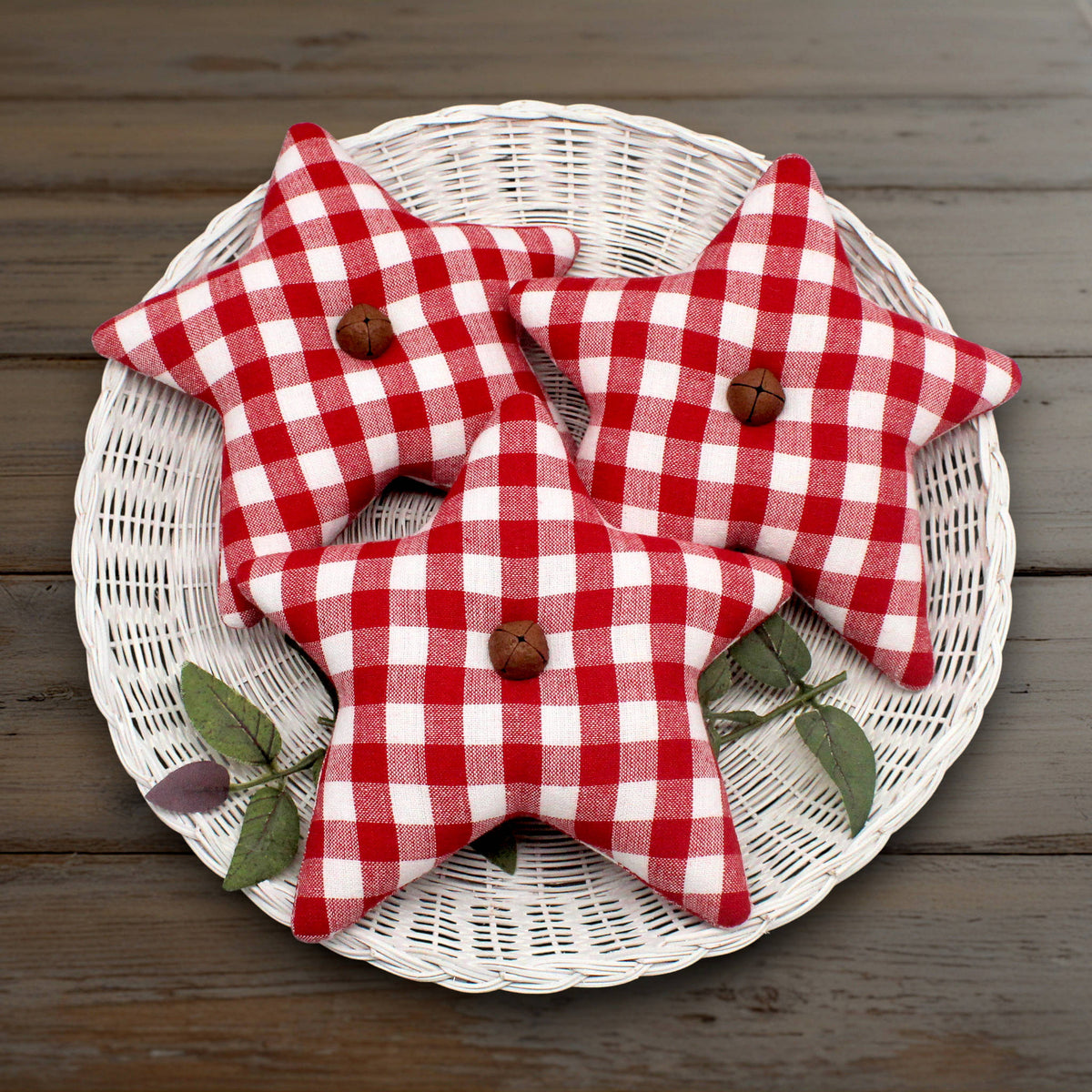 Three red and white homespun star bowl fillers with rusty bells, arranged on a white wicker plate over a dark wood surface.