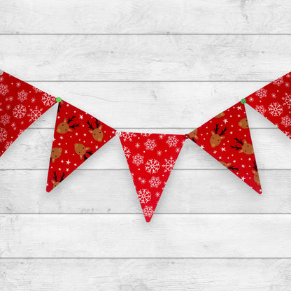 Red Christmas bunting banner with triangular flags featuring white snowflakes, brown reindeer, and small green accents, displayed against a white wood plank background.
