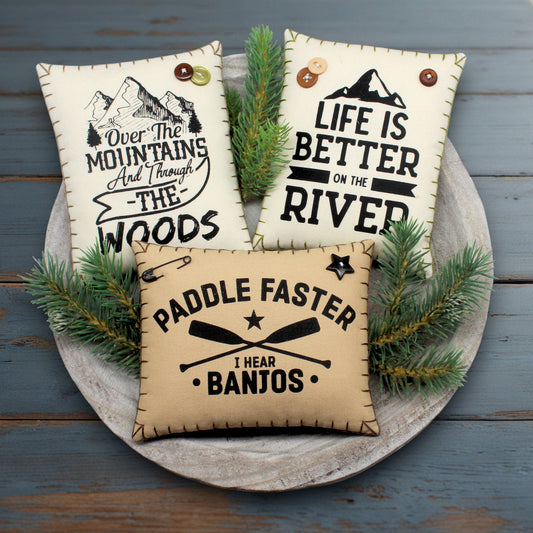 Three decorative pillows with motivational phrases about living in the mountains near the river on a wooden surface.