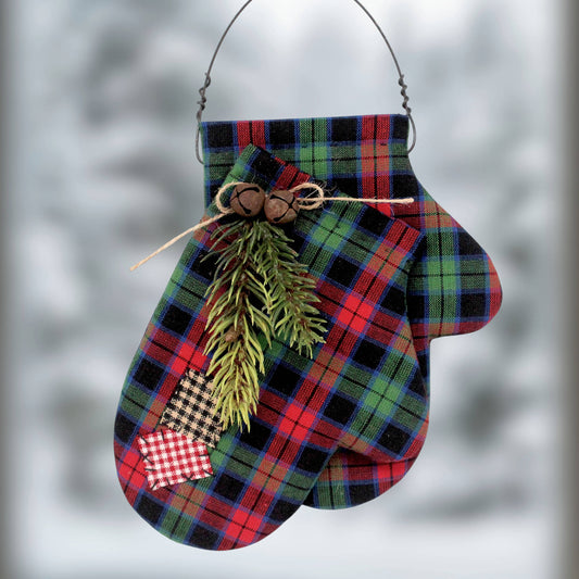 A Christmas mitten ornament made from red, green, and black plaid fabric, decorated with a pine sprig, bunting, and a small patch, hanging from a wire loop against a blurred winter background.