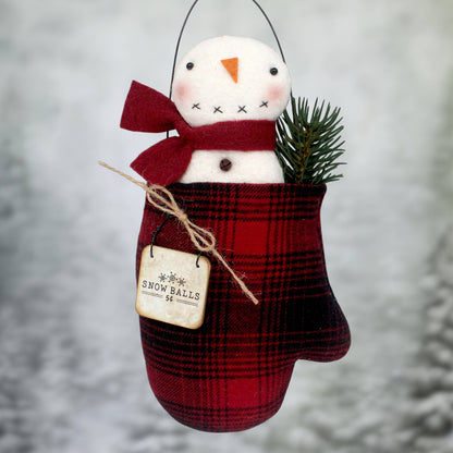 A Christmas ornament made with a primitive snowman tucked inside a black and red homespun mitten with a wire hanger and a small sign that reads 'snowballs'