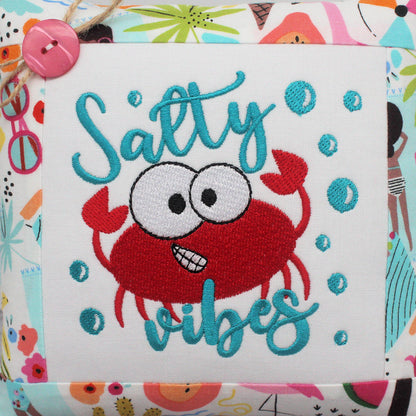 Closeup of a small square decorative throw pillow with a colorful beach-themed border and a central white panel featuring a red embroidered crab and the words 'Salty vibes' in teal script, shown against a blurred beach background.