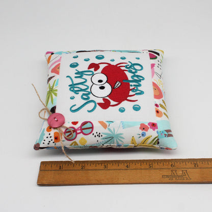 A square fabric pincushion with a colorful cartoon crab design and the words 'salty vibes' in teal script, shown on a white background with a wooden ruler below for scale.