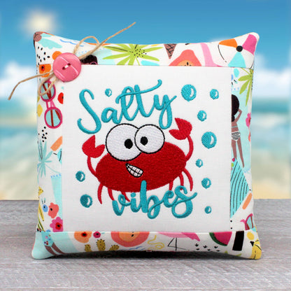 A small square decorative throw pillow with a colorful beach-themed border and a central white panel featuring a red embroidered crab and the words 'Salty vibes' in teal script, shown against a blurred beach background.