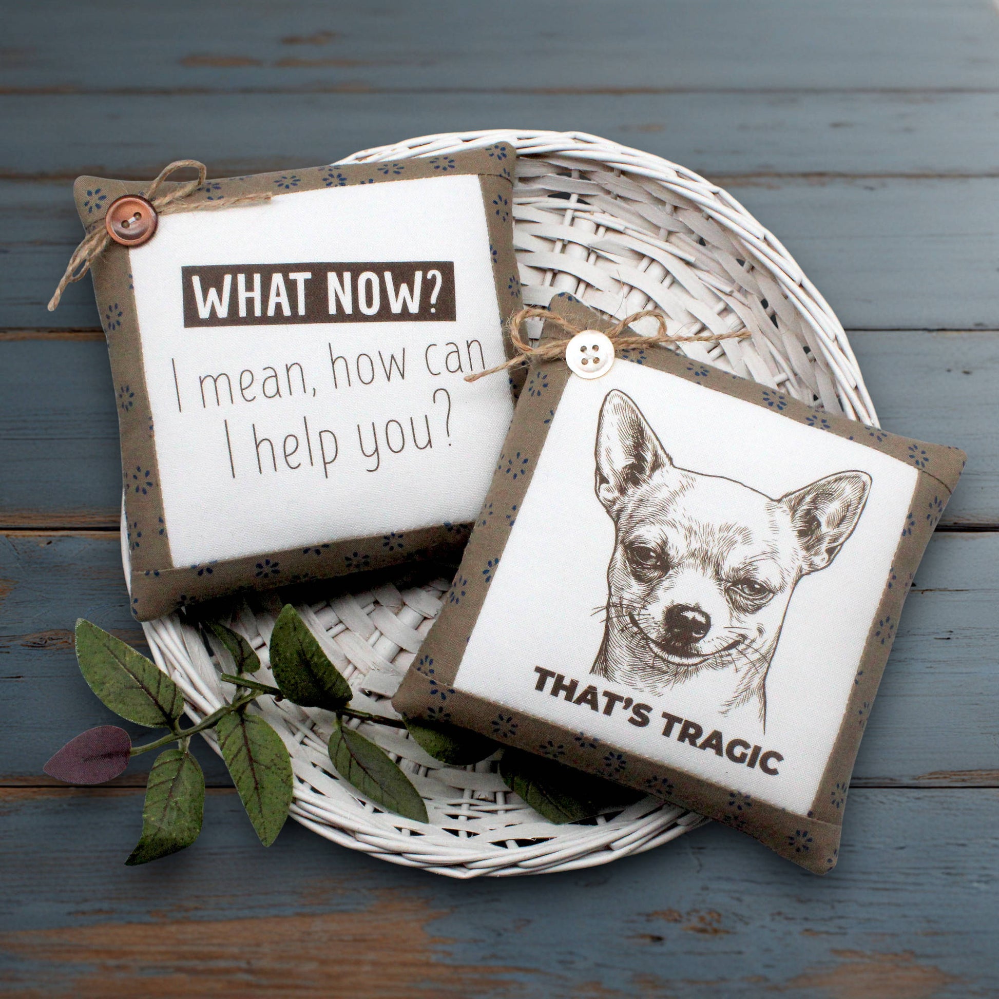 Two decorative square mini pillows displayed on a white woven tray over a distressed blue wooden surface. One pillow has a brown band with the text 'WHAT NOW? I mean, how can I help you?' and the other has a brown band with a black-and-white chihuahua illustration and the text 'THAT'S TRAGIC'.