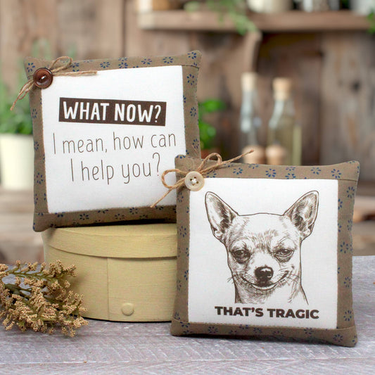 Two decorative square mini pillows with fabric trim and twine bows are displayed on a table. One pillow shows the text 'WHAT NOW? I mean, how can I help you?' and the other shows a black-and-white chihuahua illustration with the text 'THAT'S TRAGIC'.