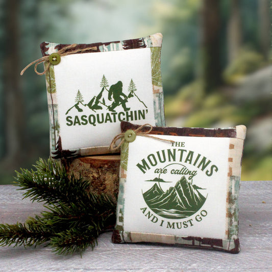 Two small square fabric pillow-style ornaments with rustic camouflage trim, displayed on a log outdoors. One reads “SASQUATCHIN” with a green sasquatch graphic, and the other reads “THE MOUNTAINS are calling AND I MUST GO” with a mountain graphic.