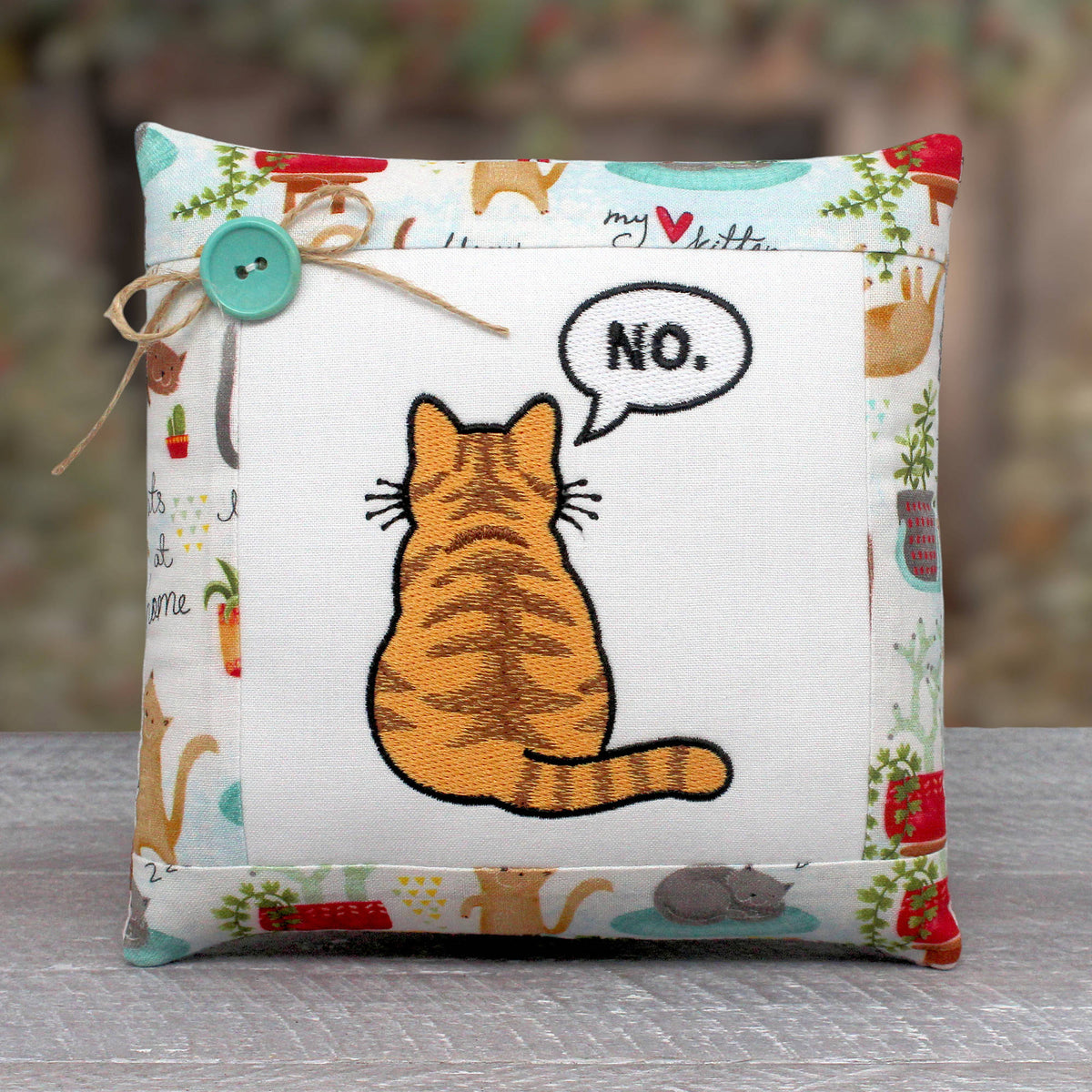 Small pillow tuck or bowl filler embroidered with a cat who has his back turned to the view with a floating speech bubble with the word 'no.'  The center panel is trimmed with a fabric with a cat design.