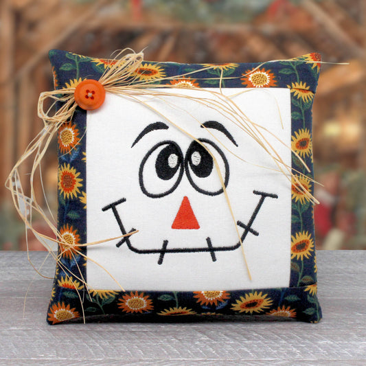 Small pillow tuck or bowl filler embroidered with a smiling scarecrow face.  The center panel is trimmed with a sunflower print fabric and there's a raffia bow tied around a button at the top corner.