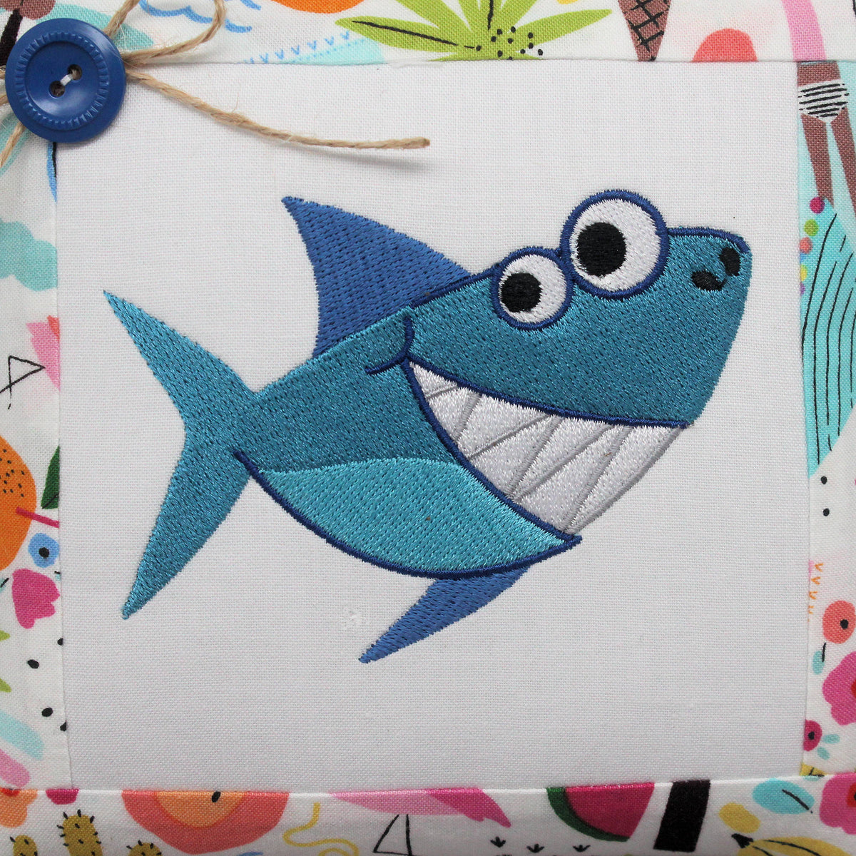 A close-up of a blue embroidered cartoon shark design on white fabric, with a colorful patterned border around the edges and a small blue button with twine in the top-left corner.