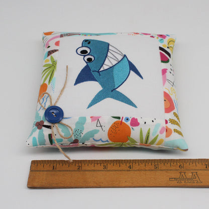 A square fabric pillow with a colorful tropical print border and a blue embroidered shark on the front, shown next to a wooden ruler for scale on a white background.