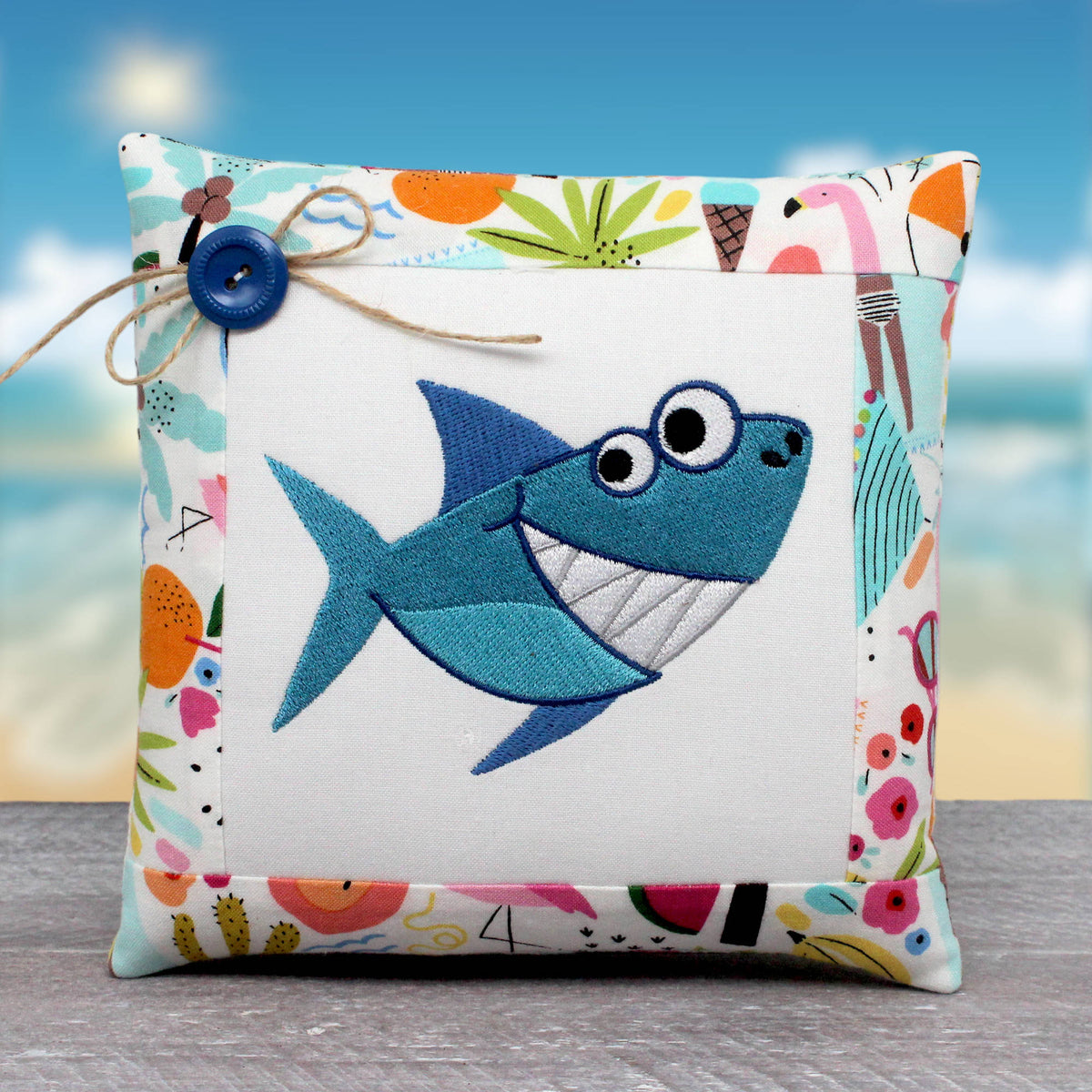 A square decorative pillow with a colorful patchwork border and a central embroidered blue shark on a white background, styled with a twine bow and blue button, set against a beach-themed backdrop.
