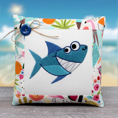 A square decorative pillow with a colorful patchwork border and a central embroidered blue shark on a white background, styled with a twine bow and blue button, set against a beach-themed backdrop.