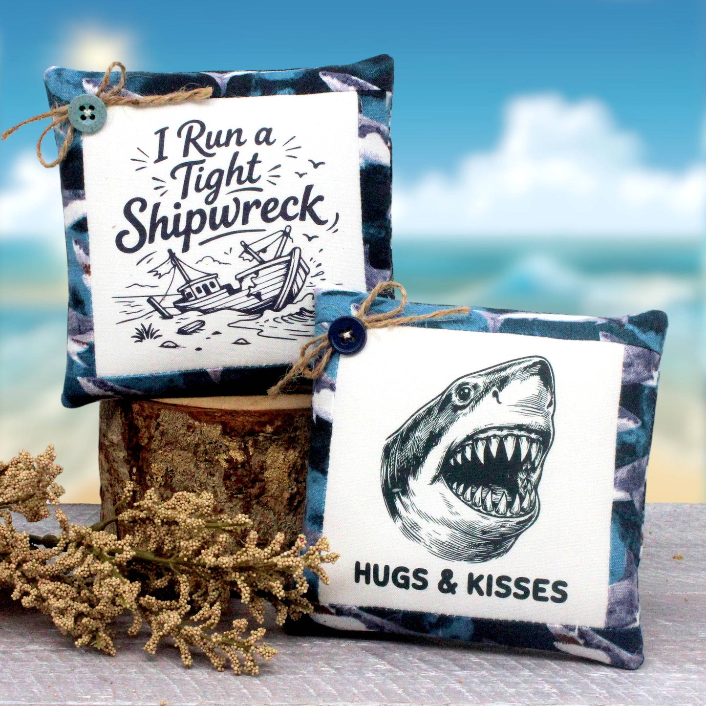 Two decorative square mini pillows with blue and white fabric, styled on a rustic wood slice with dried coral. One pillow features a white square panel with the text 'I Run a Tight Shipwreck' and a ship illustration, and the other features a shark face illustration with the text 'HUGS & KISSES'.
