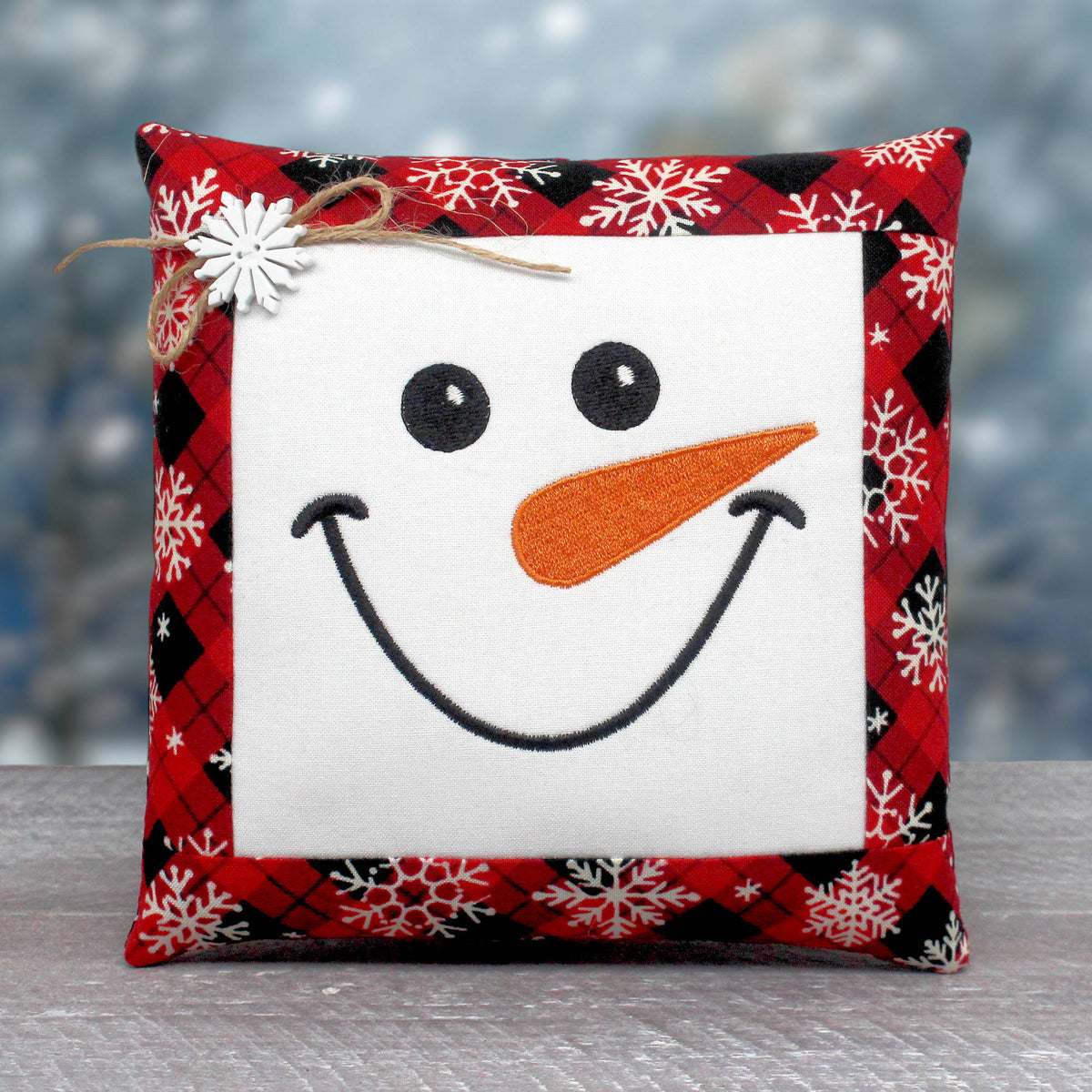A square decorative throw pillow with a snowman face design. The center is white with black embroidered eyes and smile, an orange carrot nose, and a red, black, and white plaid border with white snowflakes.