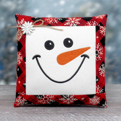 A square decorative throw pillow with a snowman face design. The center is white with black embroidered eyes and smile, an orange carrot nose, and a red, black, and white plaid border with white snowflakes.