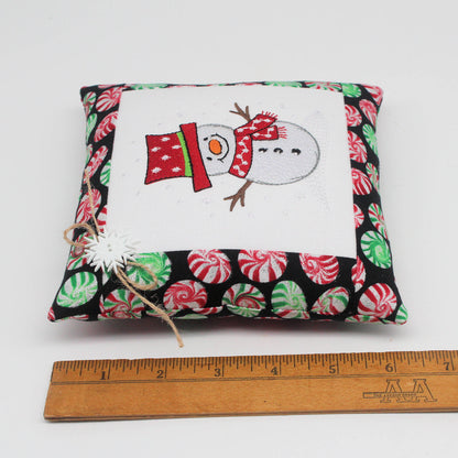 A small square fabric pillow with a Christmas snowman design and candy cane pattern, shown with a wooden ruler below for scale on a white background.